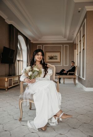 Bride holding bouquet while seated in elegant room with classic decor.