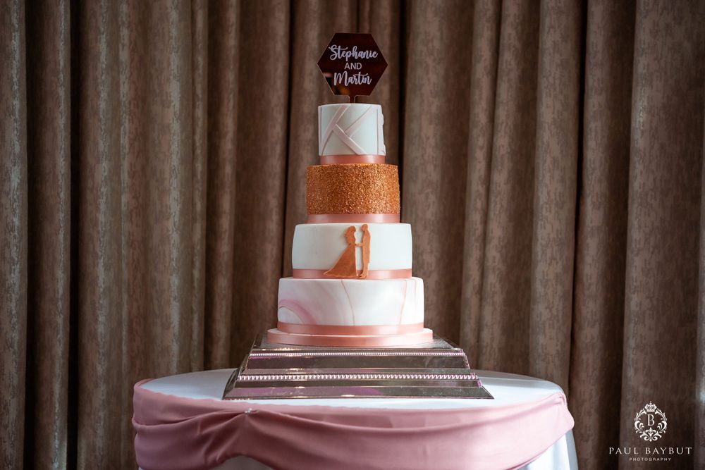 Four-tier wedding cake with rose gold and pink accents displayed on a pink tablecloth.
