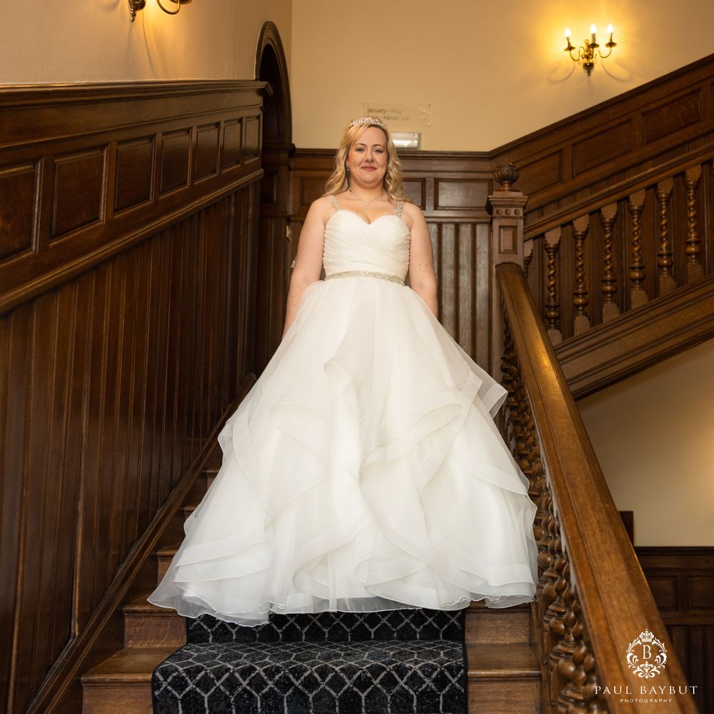 A wedding gown with a layered tulle skirt stands elegantly on a grand wooden staircase with ornate carpeting.