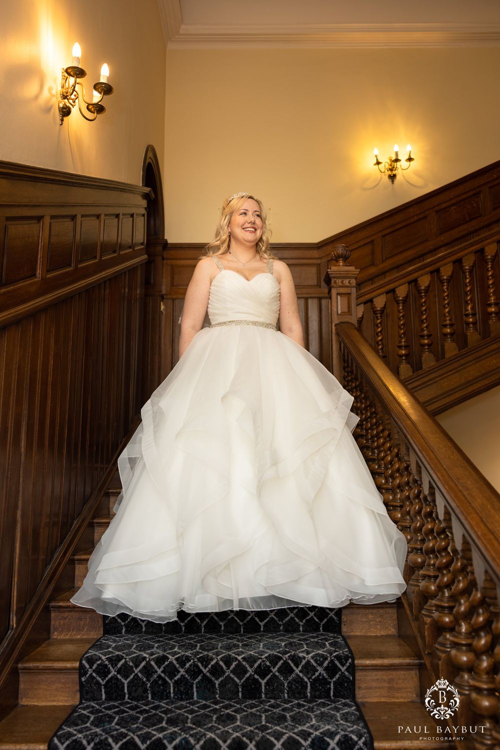 A flowing white wedding dress with ruffled layers photographed on carpeted stairs with wooden banisters.