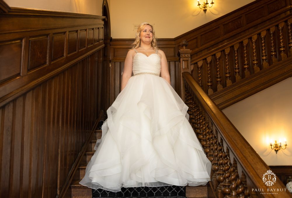 An elegant bridal gown photographed from below on a classic wooden staircase with wall sconces.