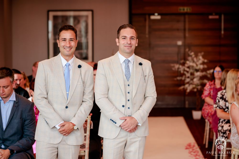 Two groomsmen in matching light beige suits with blue ties stand at the altar.