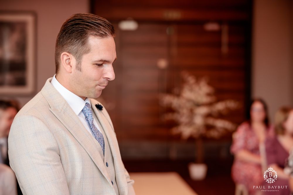 A formal wedding participant in cream colored suit and blue tie stands thoughtfully.