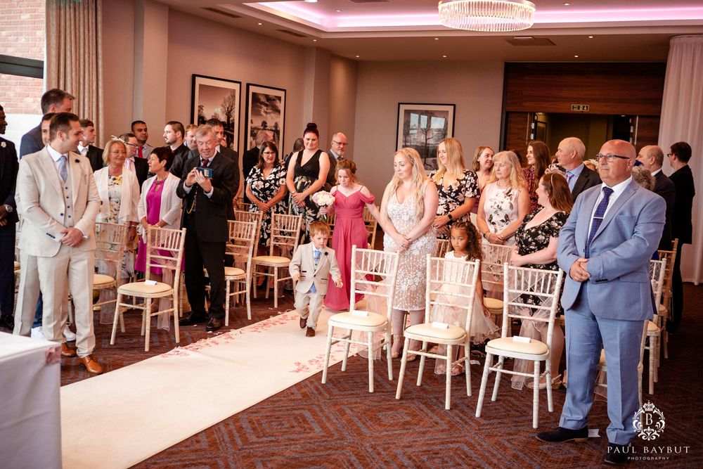 Wedding guests stand during ceremony in elegant ballroom with white chairs and pink accent lighting.