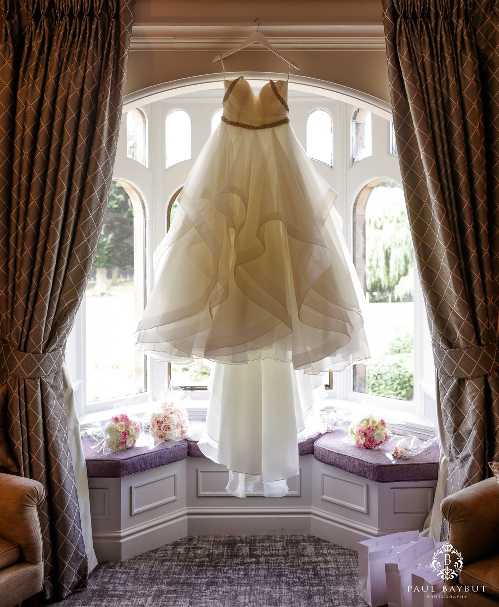 White wedding dress hanging in elegant bay window with draped curtains and decorative details.