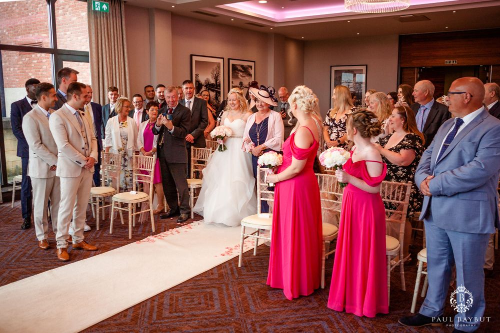 Wedding ceremony in progress with bridesmaids in pink dresses and white aisle runner.