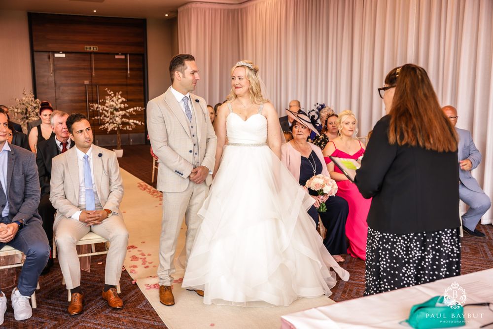 Wedding ceremony underway in elegant ballroom with white decor and pink lighting.