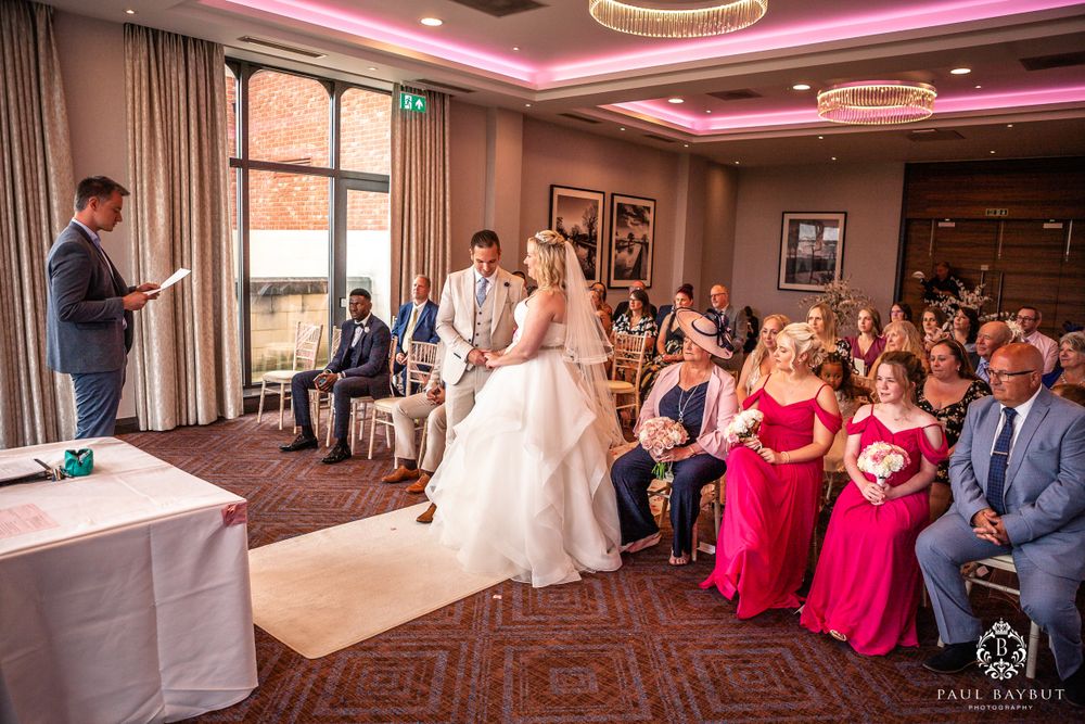 Wedding ceremony in an elegant ballroom with pink lighting and rows of seated guests watching.