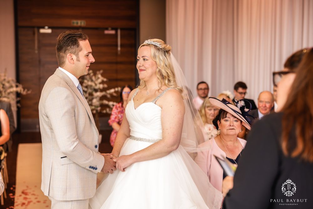 Intimate exchange of vows during indoor wedding ceremony with soft lighting and curtained backdrop.