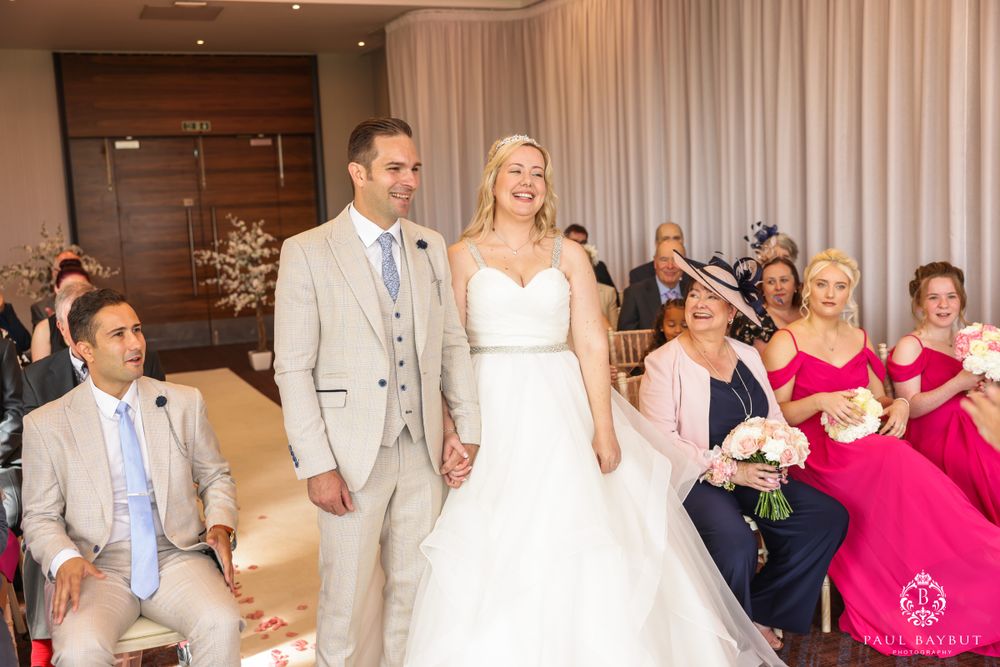 Wedding couple shares a joyful moment during their ceremony with bridesmaids in pink dresses looking on.
