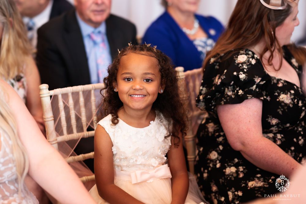 Smiling young attendee in white dress sits among wedding guests.