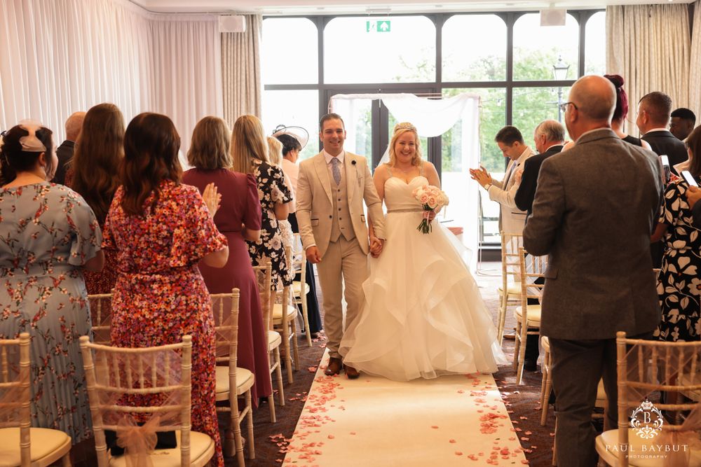 Couple stands at end of aisle during wedding ceremony with guests seated in decorated venue.
