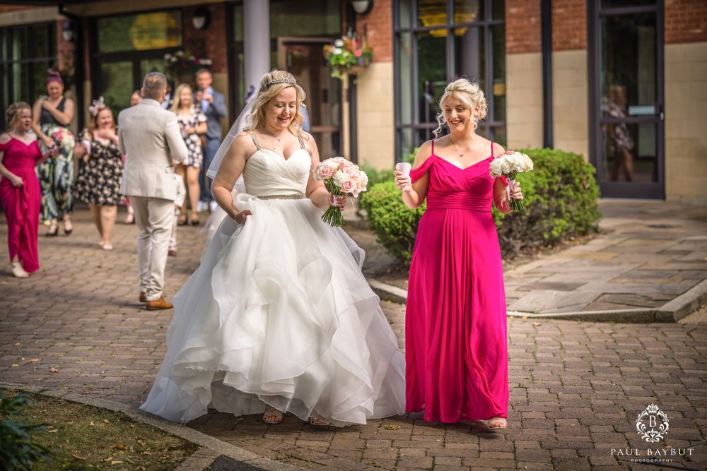 Bride in white gown walks outdoors with bridesmaid in bright pink dress.