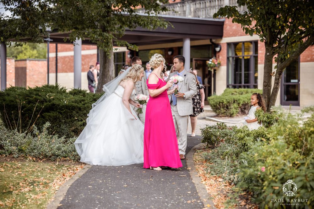 Wedding party walking along garden path with person in white dress and bright pink dress at a brick venue.
