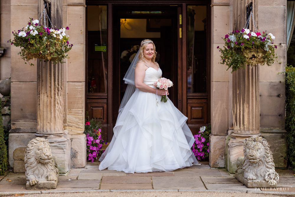 A bride in a white ballgown poses in the elegant stone doorway of a historic building flanked by hanging floral baskets.