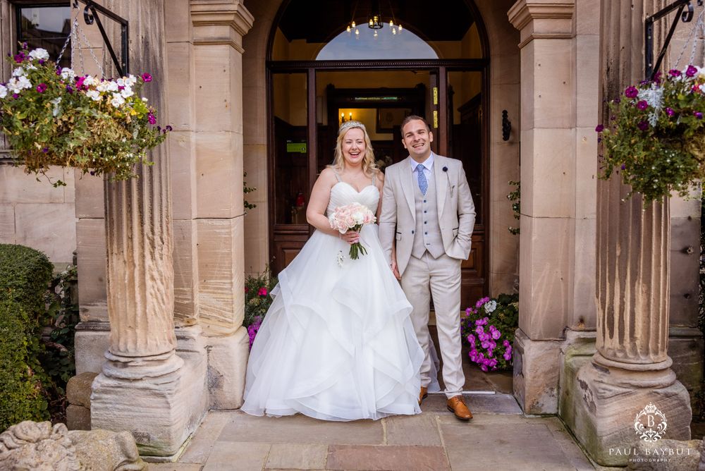 The newlyweds stand together in a classic stone entryway adorned with hanging floral arrangements.