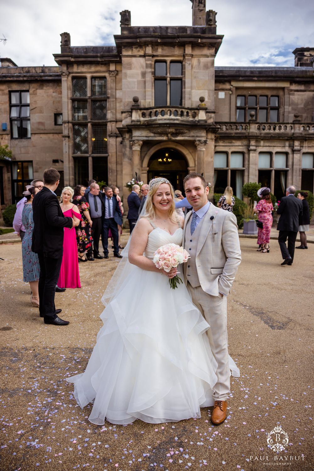 A wedding portrait in front of a historic stone manor with guests and confetti in the background.