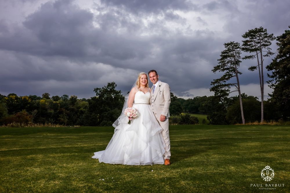 A wedding photo of a couple on a grassy lawn with dramatic storm clouds and tall pine trees in the background.