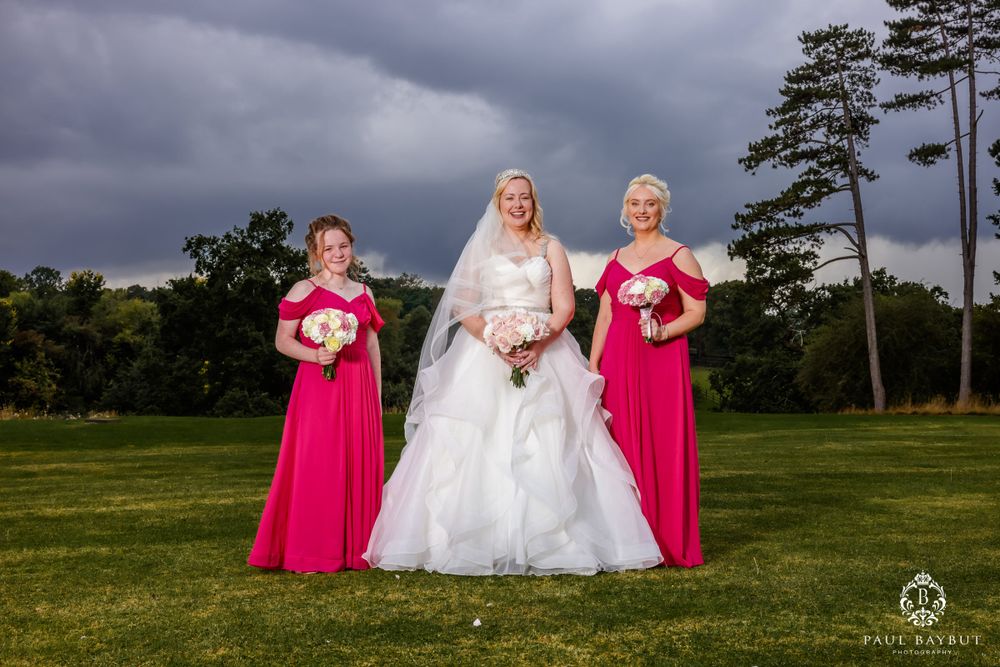 Wedding party members in bright pink dresses pose with bride on a lawn under stormy skies.