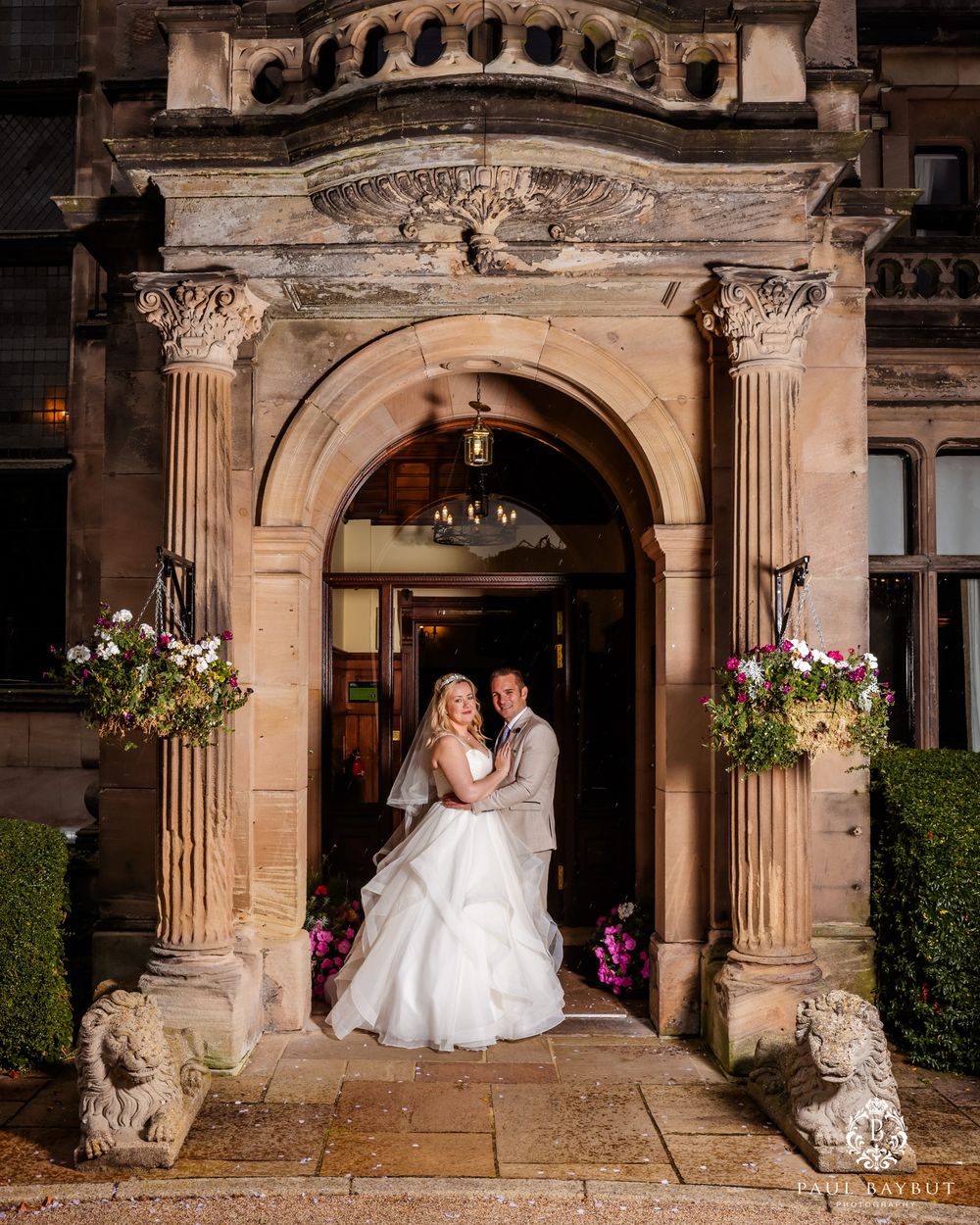 Wedding couple poses in an ornate stone archway entrance with hanging flower baskets.