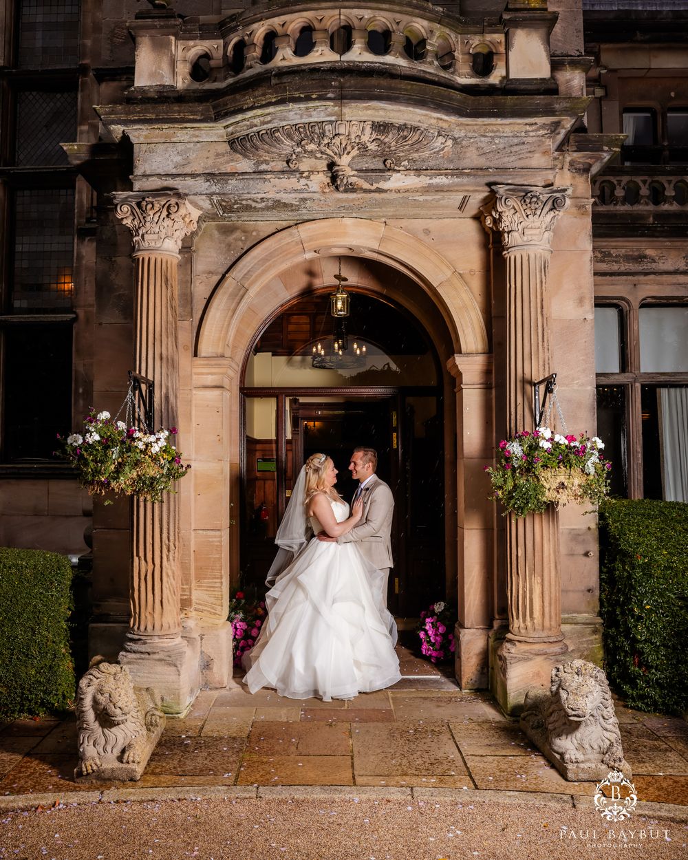 Romantic evening portrait of newlyweds under a decorative stone archway with floral arrangements.