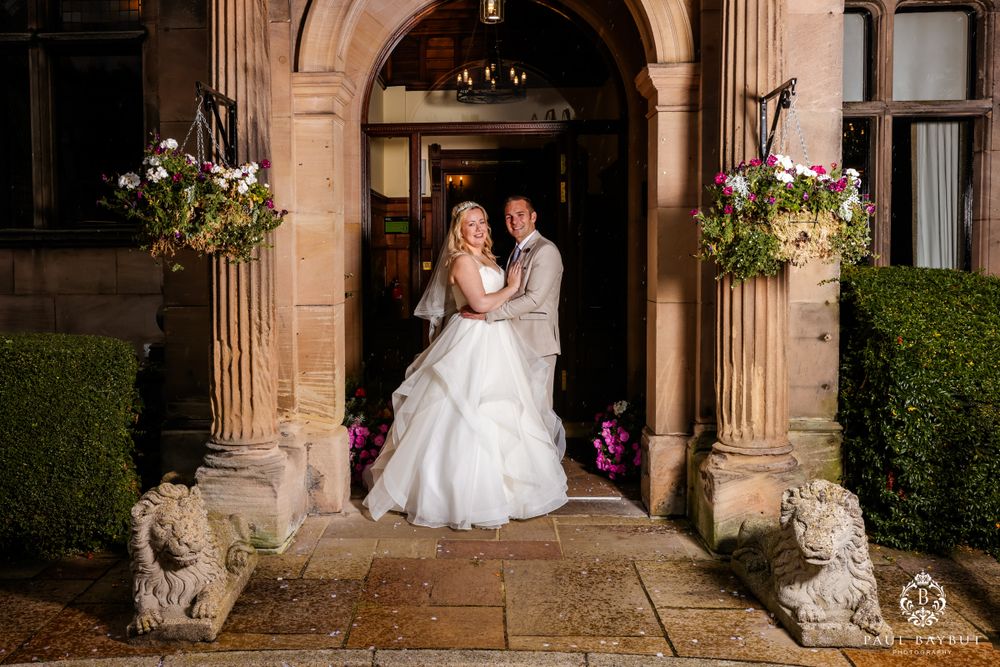 A wedding photo taken at a stone archway entrance framed by decorative stone lions and floral arrangements.