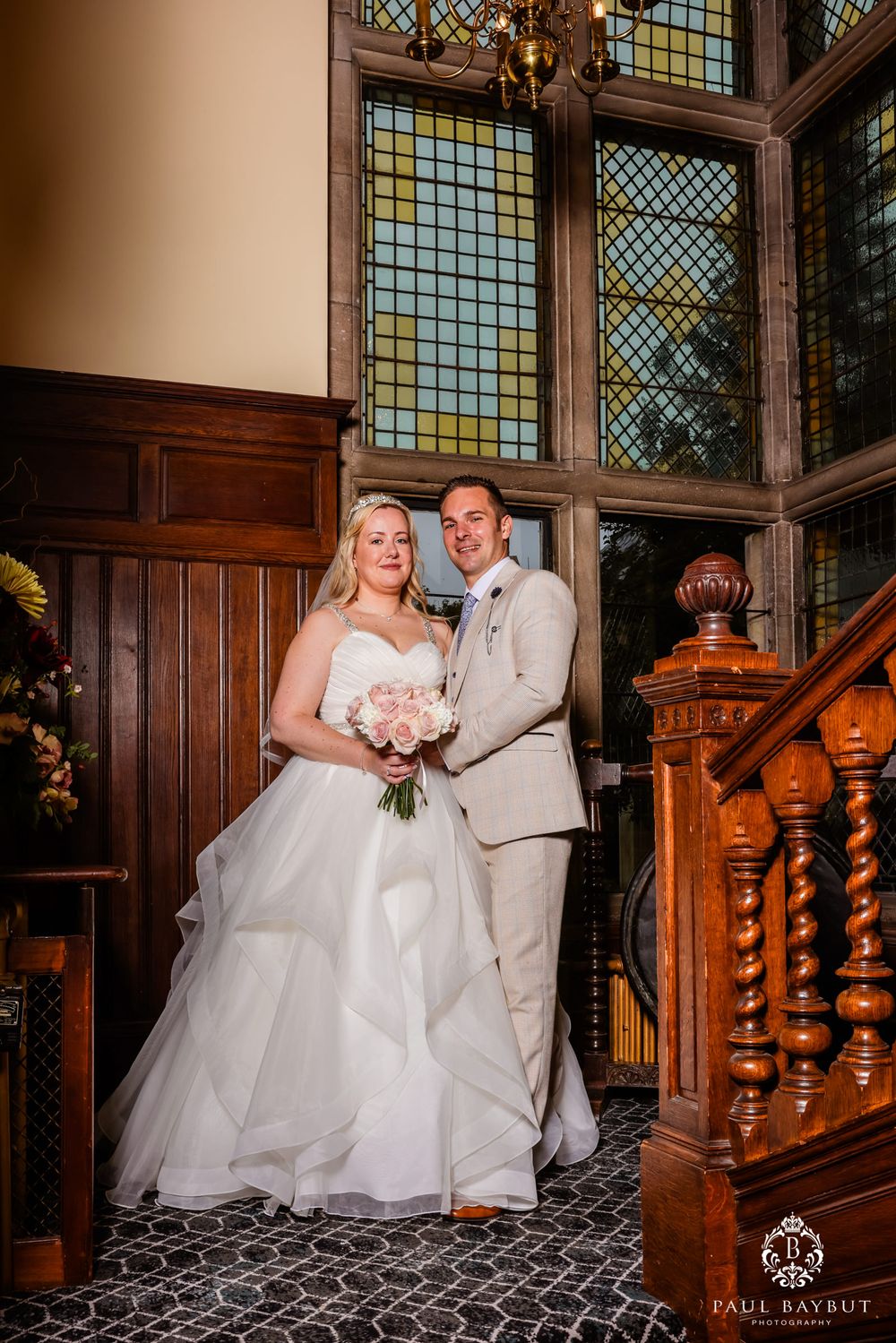 An elegant wedding portrait featuring a newlywed couple standing by a vintage wooden staircase and stained glass windows.
