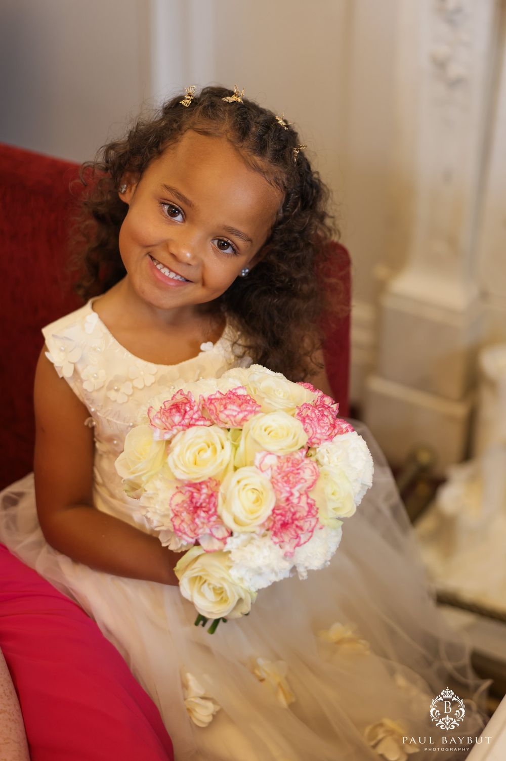 A young flower girl in a white dress smiling while holding a bouquet of pink and white roses.