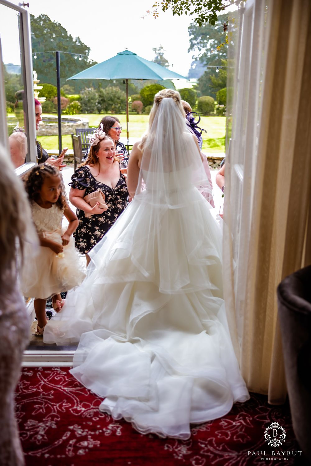 A bride in a flowing white gown walking on red carpet with attendants and guests visible in garden setting.
