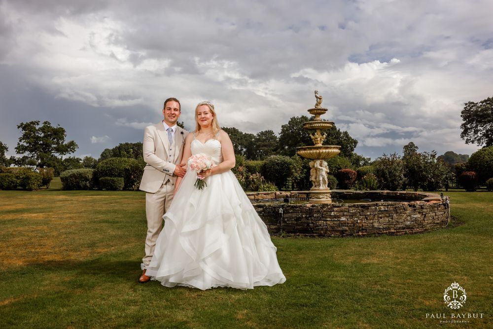 Newlyweds pose together in garden setting with ornate fountain and dramatic cloudy sky.