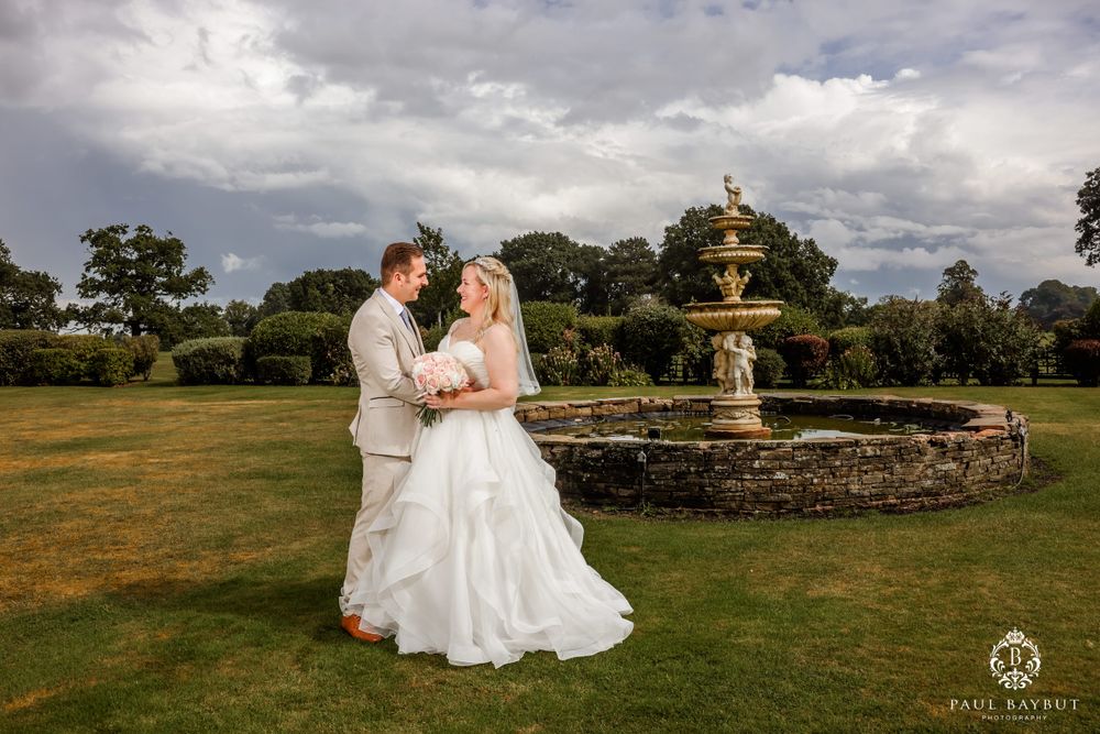 Romantic wedding couple embraces by stone fountain under dramatic afternoon sky.
