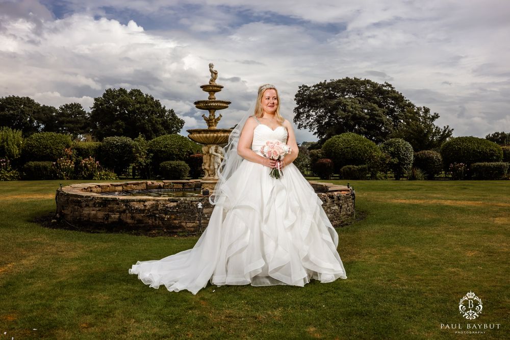 Elegant bride in flowing white gown poses beside ornate garden fountain under cloudy skies.