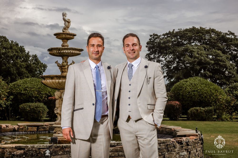 Groomsmen in light grey suits smile together by ornate garden fountain.