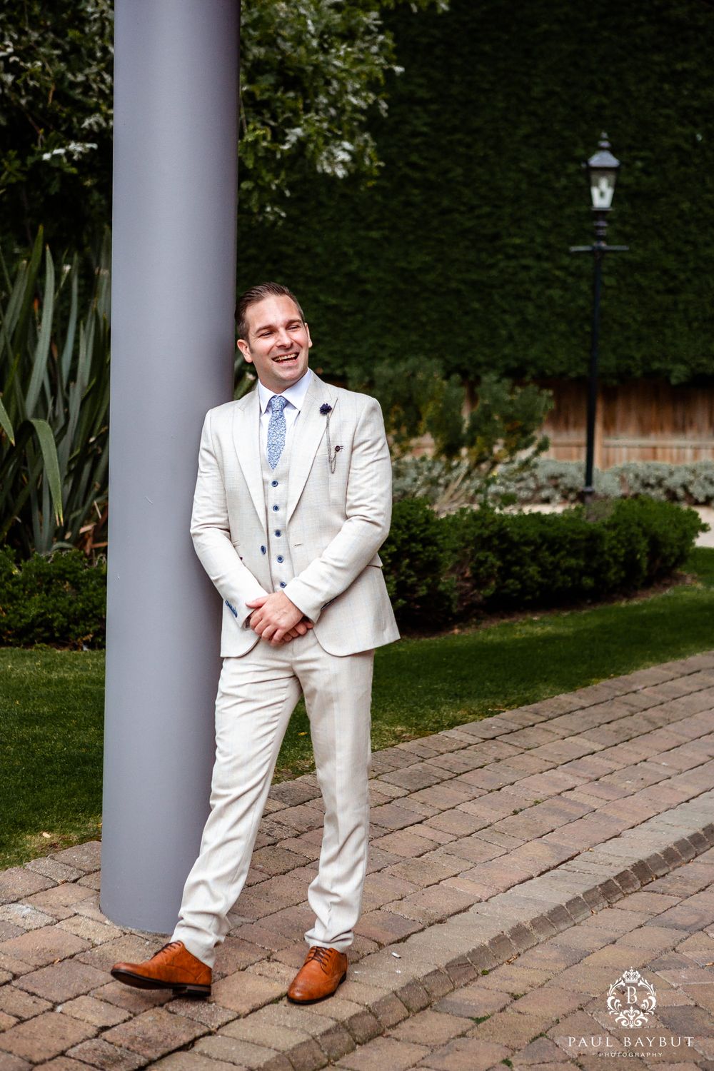 Person in light colored wedding suit standing by column in manicured garden setting.
