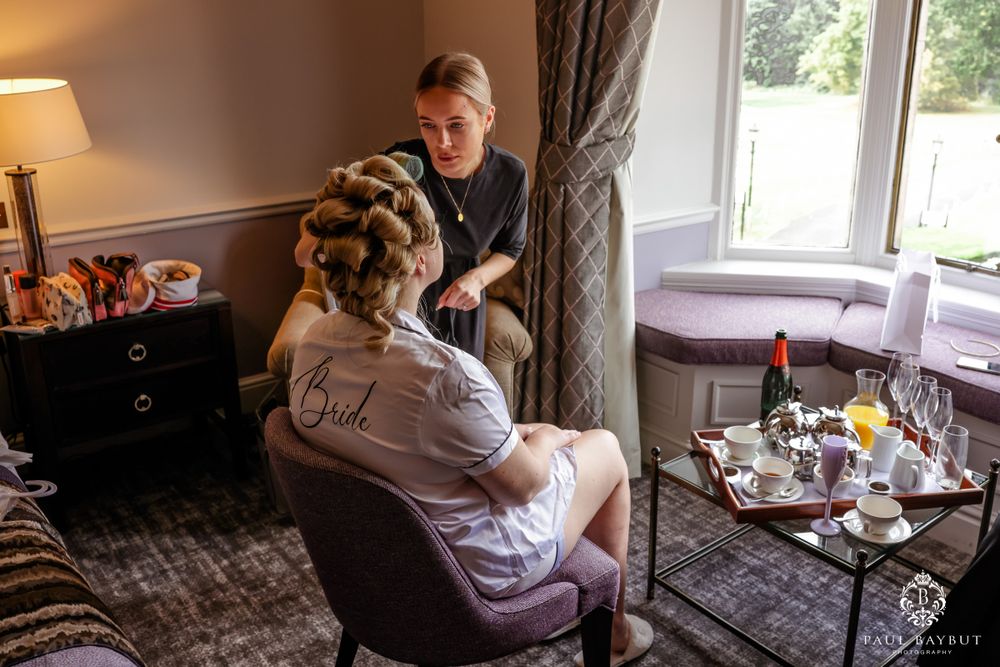 Wedding preparation scene in a hotel room with stylists working on hair and makeup.