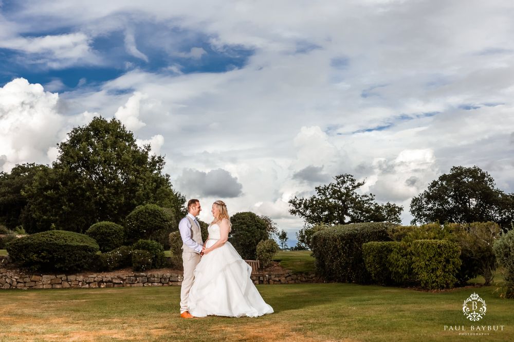 A wedding couple stands in a scenic garden setting with dramatic cloud formations overhead.