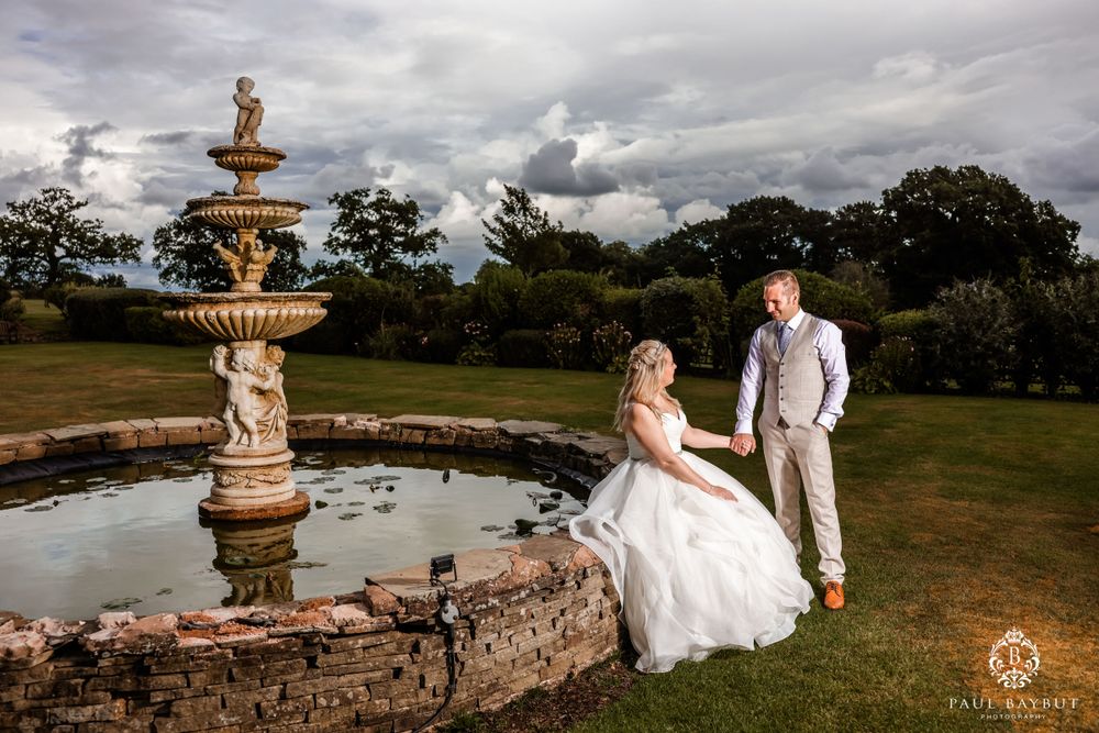 Couple poses by ornate garden fountain under dramatic cloudy sky in elegant outdoor wedding setting.