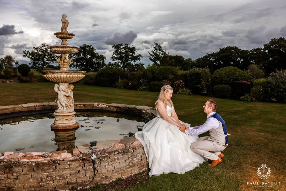 Romantic wedding scene with couple sitting by decorative stone fountain at sunset.