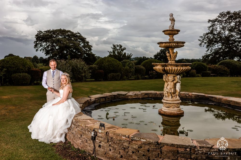 Elegant garden wedding photo with stone fountain and dramatic sky backdrop.