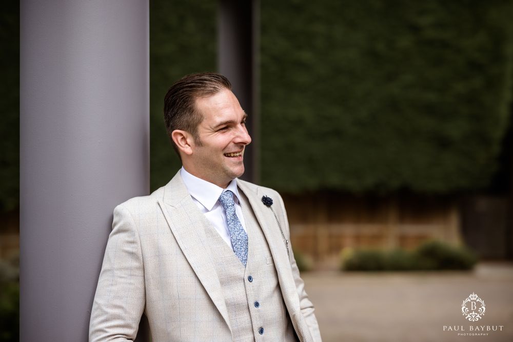 Person in three-piece light gray suit and blue tie smiling outdoors by pillar.