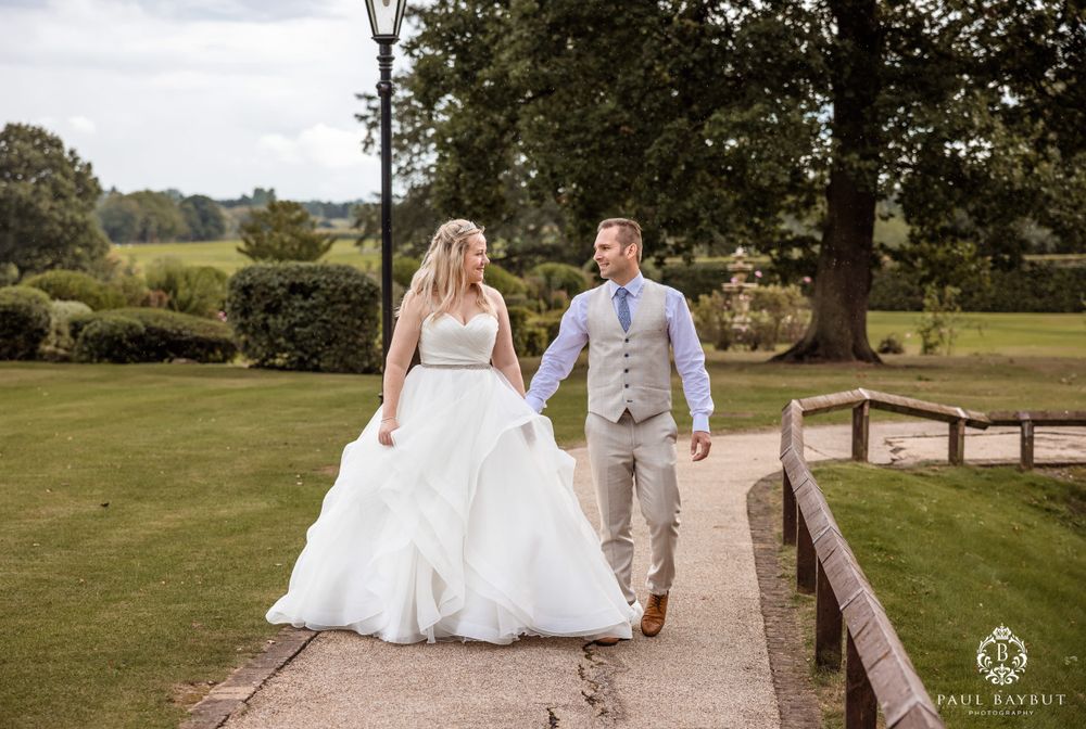 Couple walking along wooden bridge pathway through manicured garden grounds.