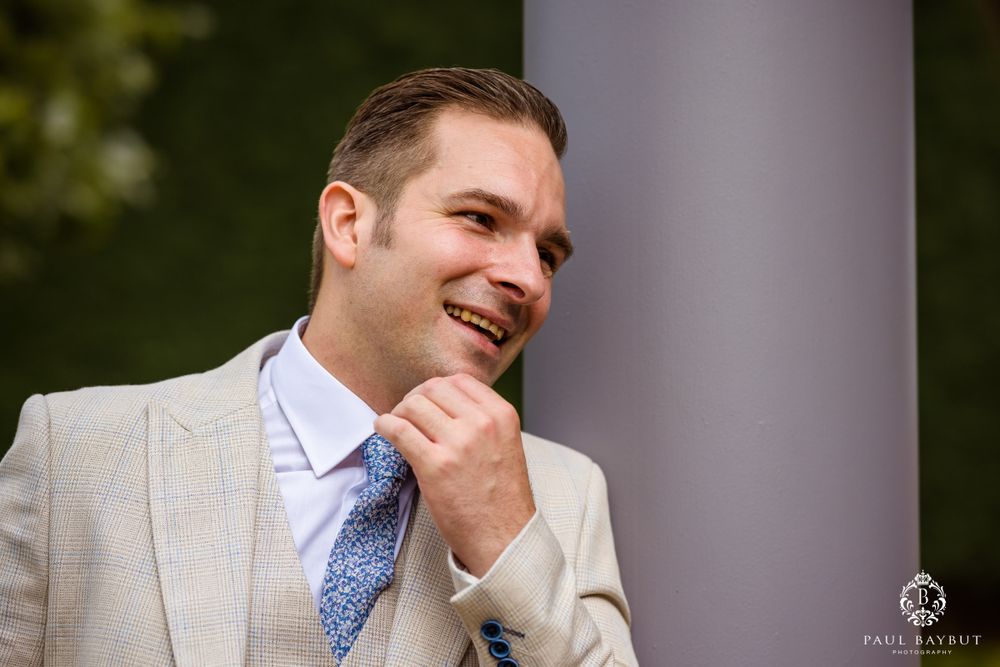 Person in light gray suit and blue tie poses by column with joyful expression.