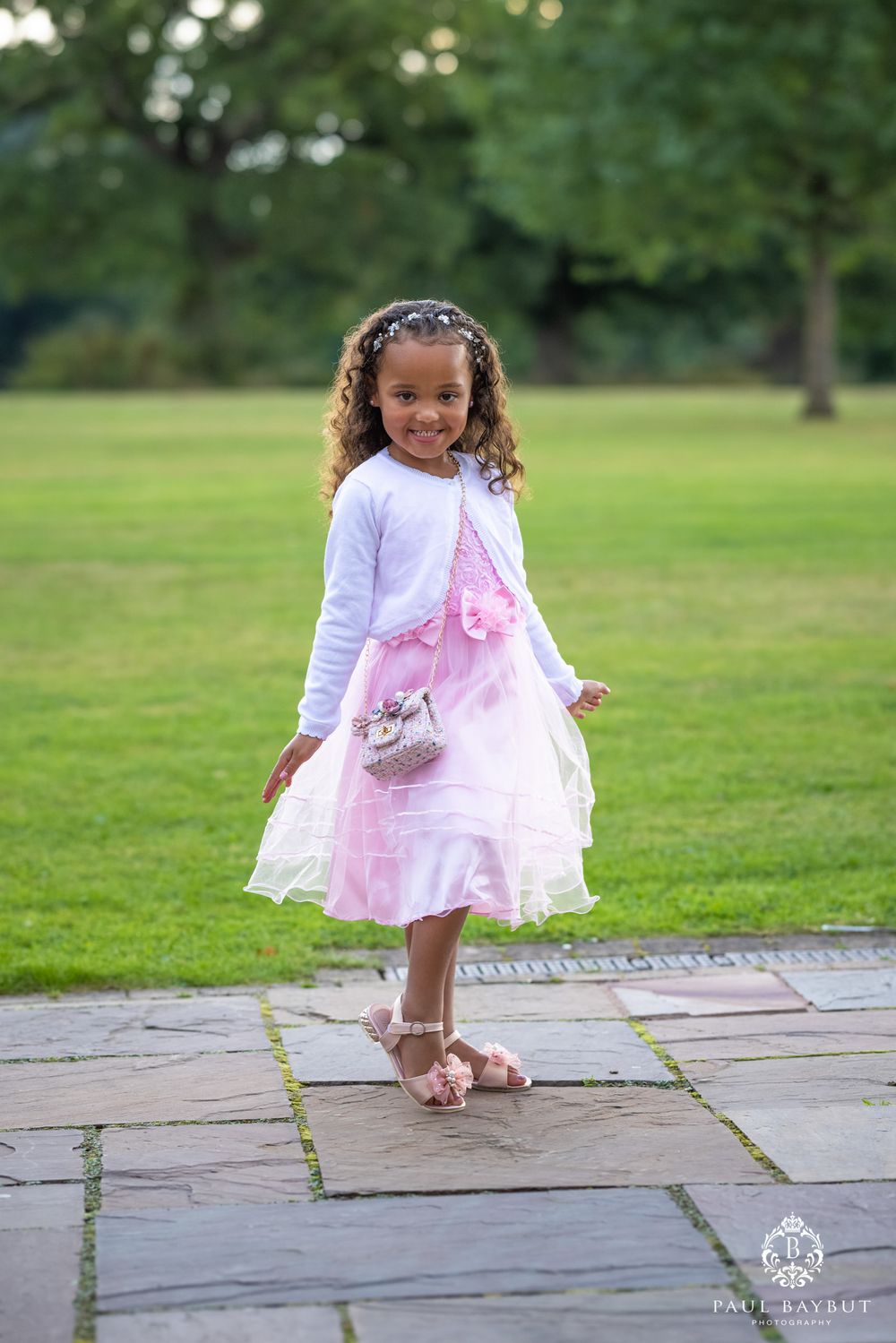 A young flower girl in a pink and white dress poses playfully on a stone path in a garden.