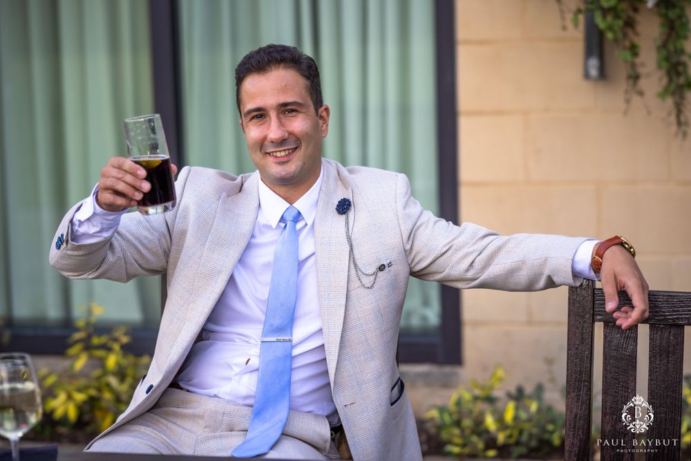 Wedding guest in light gray suit raises a glass while sitting outdoors.