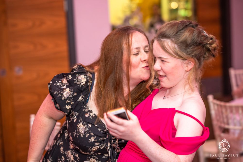 Two wedding guests share an intimate moment at a reception with pink lighting.