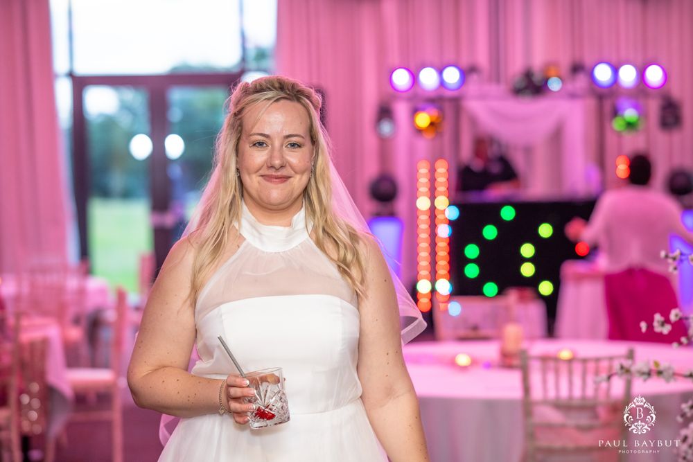 A bride stands in a wedding reception venue decorated with pink lighting and white chairs.