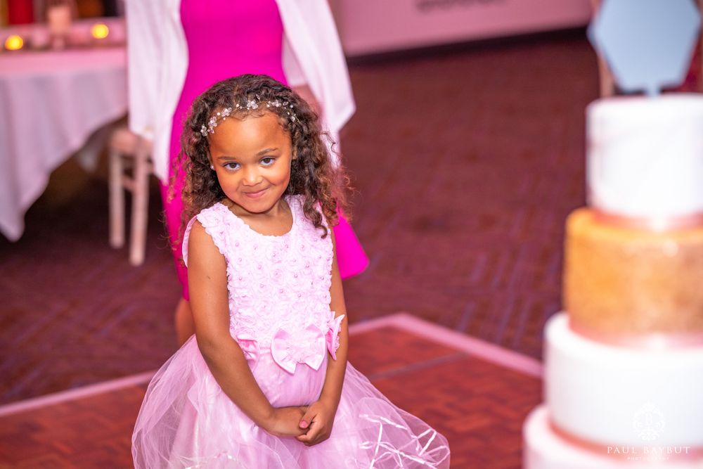 A young girl in a pink dress smiles at a wedding reception with a cake in the background.
