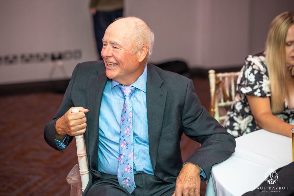 A senior gentleman in a suit and blue tie laughs while seated at an event.