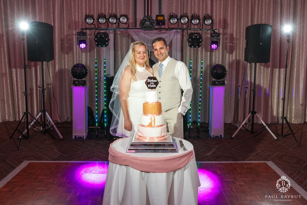 Newlyweds pose with their wedding cake in front of purple-lit DJ equipment.