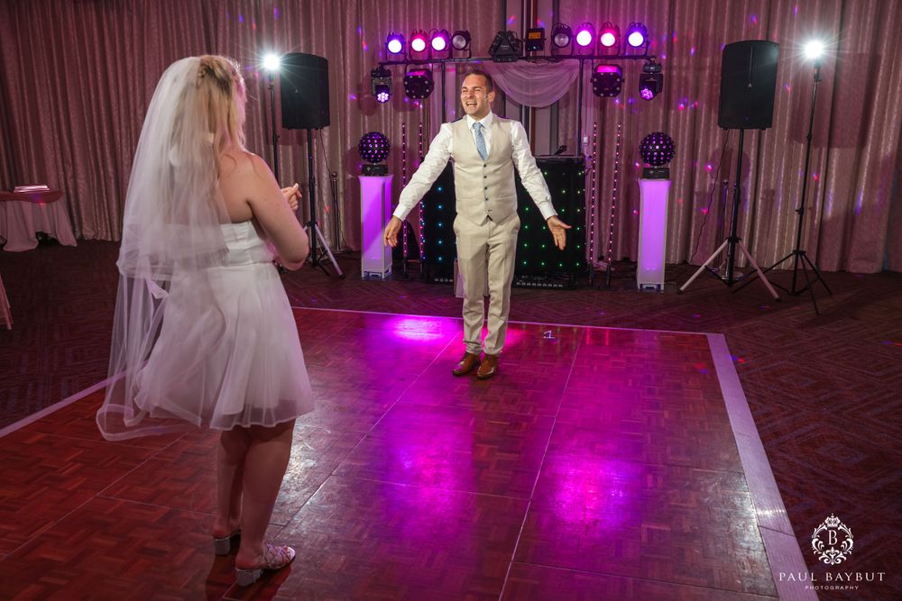 Two people dance playfully on a wooden dance floor with purple uplighting and DJ equipment.